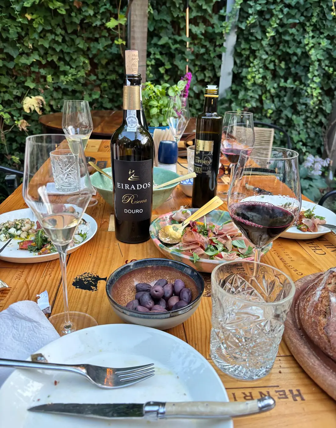 Een gedekte tuintafel met een geopende fles Eirados Reserva, presunto, olijven, brood en glazen rode en witte wijn.
